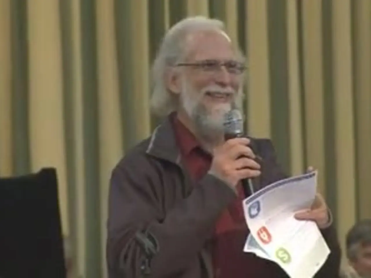 In the center of the image there is a person holding a mic and paper. In the background we can see...