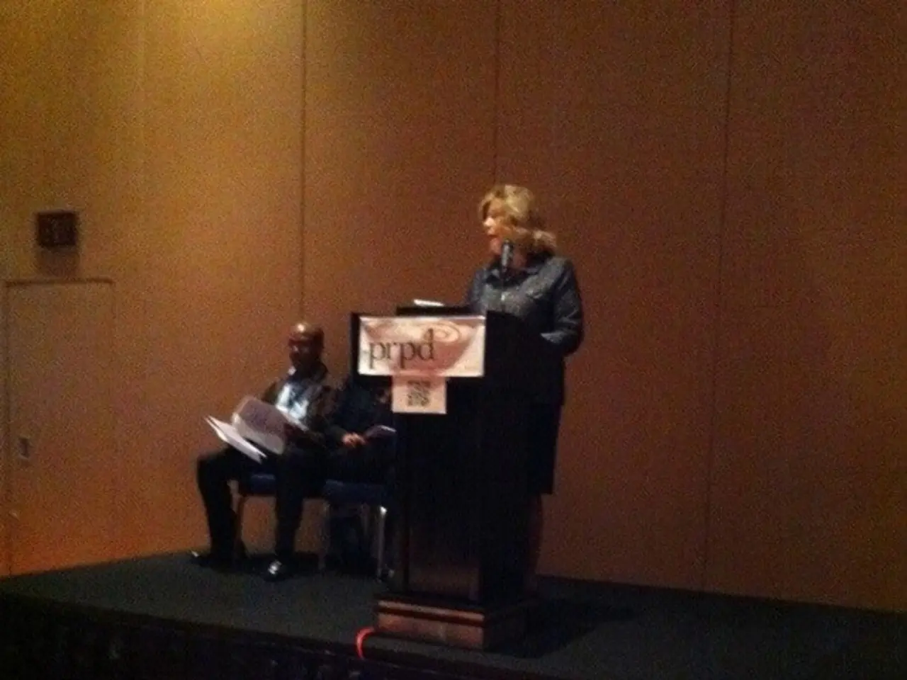 In this image there are two men sitting on chairs and woman standing in front a podium on a stage,...