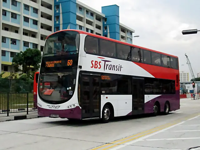 In this image there is an electric bus in the middle. In the background there is a building. On the...