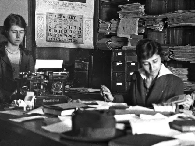 As we can see in the image there is a wall, poster, papers, two people sitting on chairs and table....